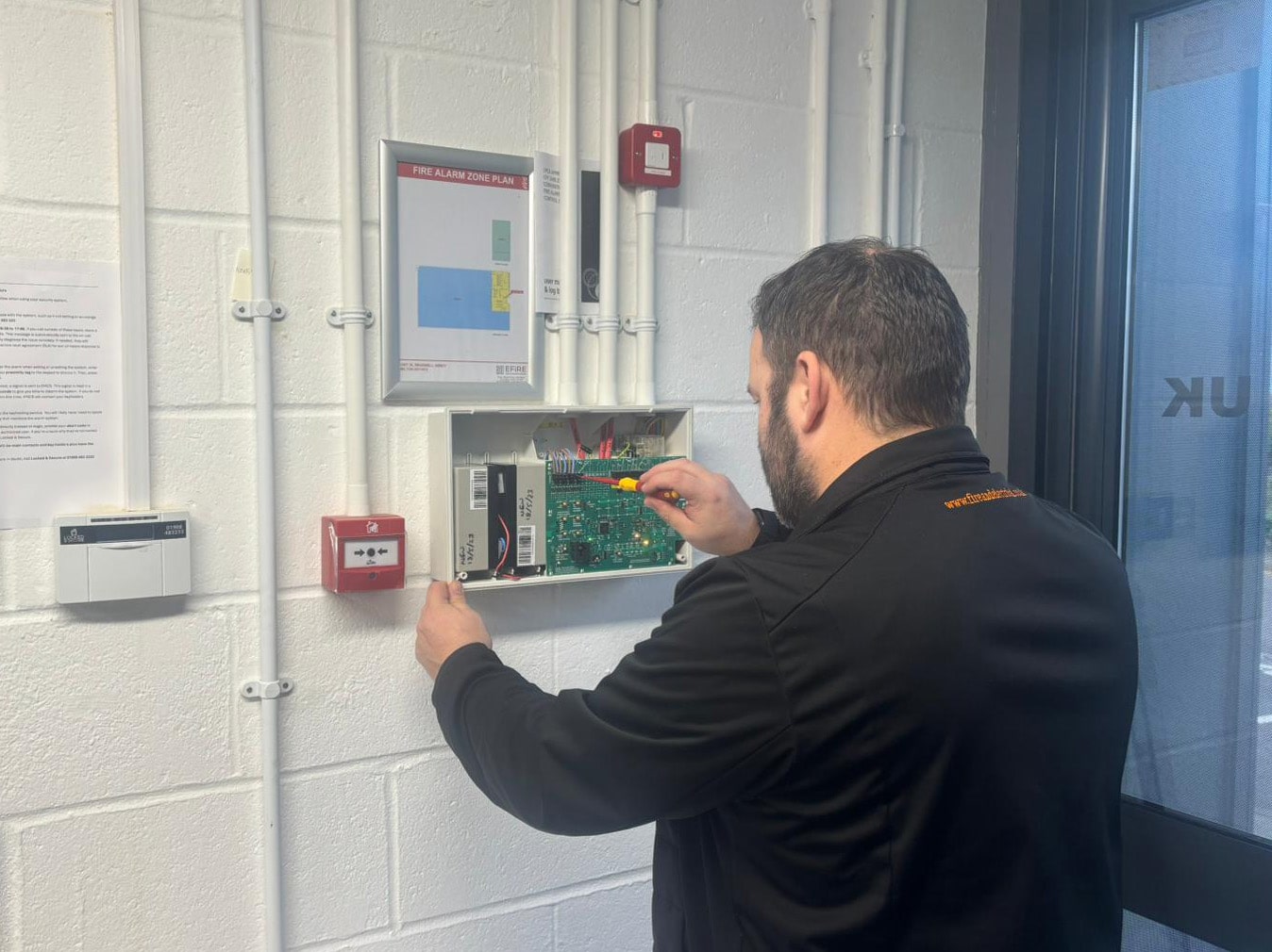 A technician inspects and repairs a fire alarm control panel on a wall.