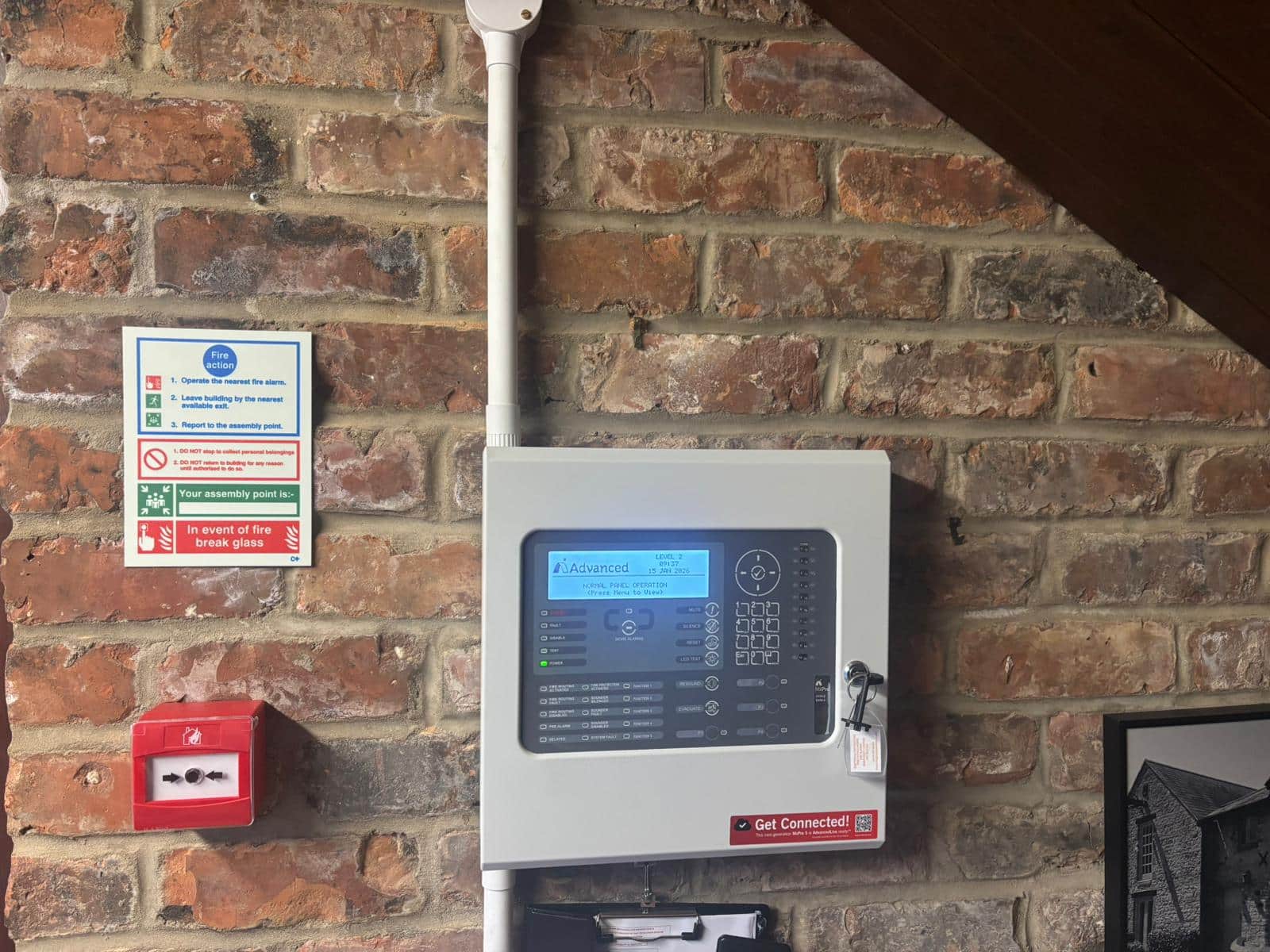 Fire alarm panel and instructions mounted on a brick wall with a red emergency break glass.
