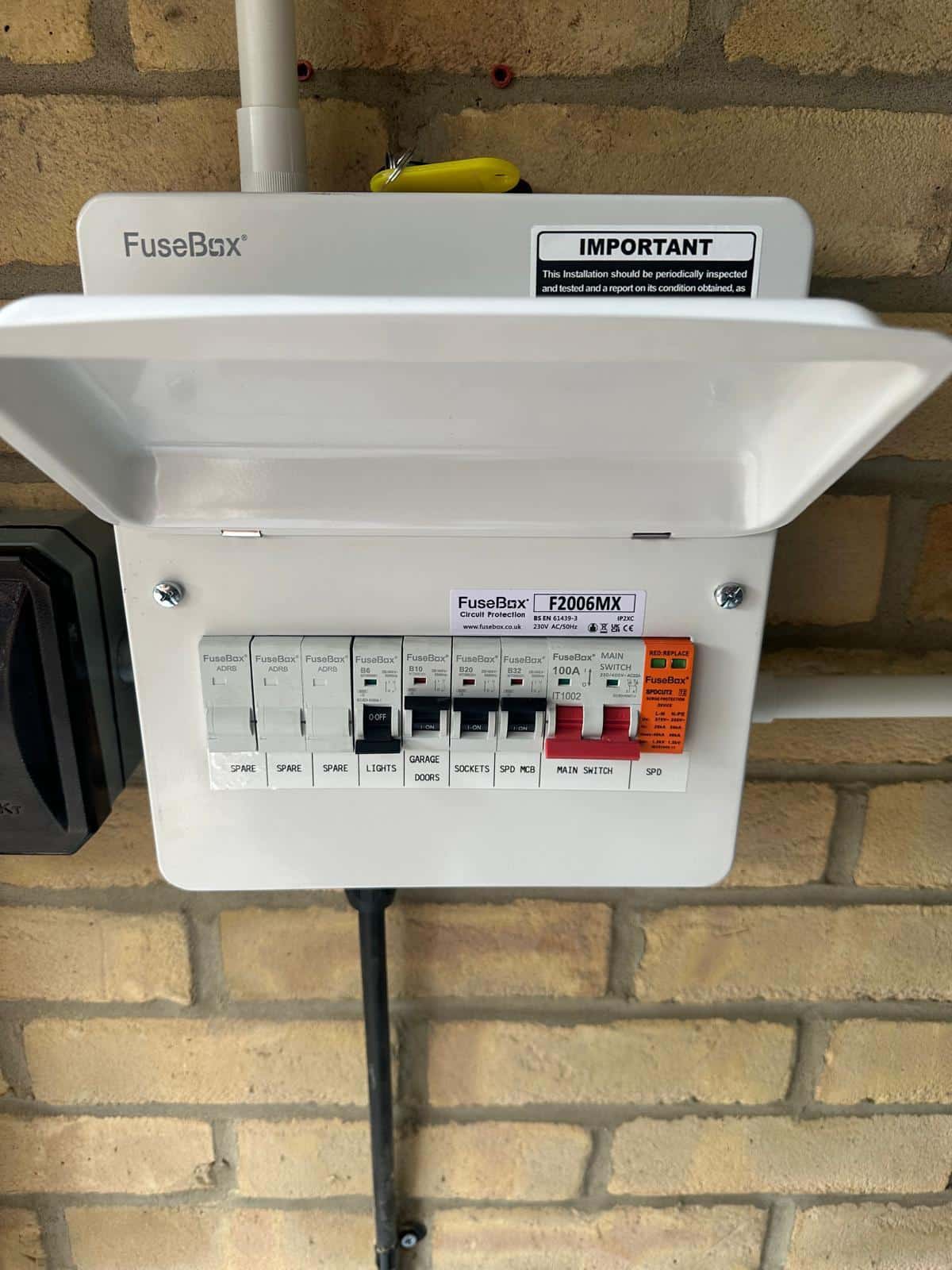 A white FuseBox electrical consumer unit with labeled switches mounted on a brick wall.