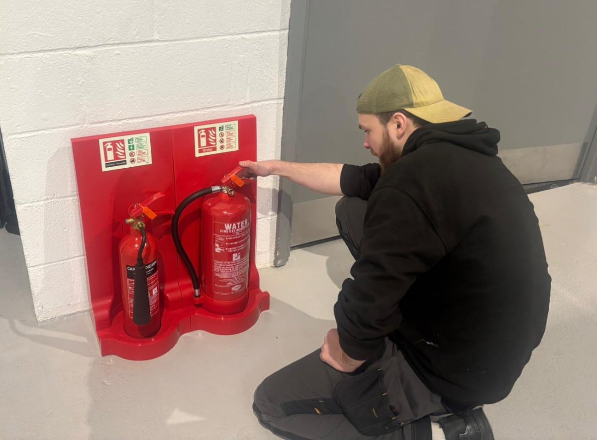 A man inspects two fire extinguishers mounted on a red stand by a wall.