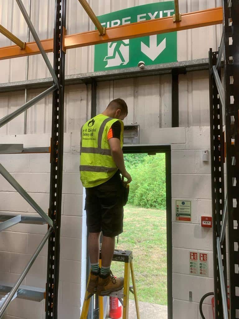 A worker on a step ladder inspects an emergency exit under a green fire exit sign.
