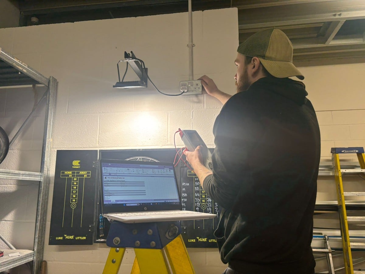 How our PAT Testing Works A man performs electrical testing on a wall outlet with a laptop on a stepladder nearby.