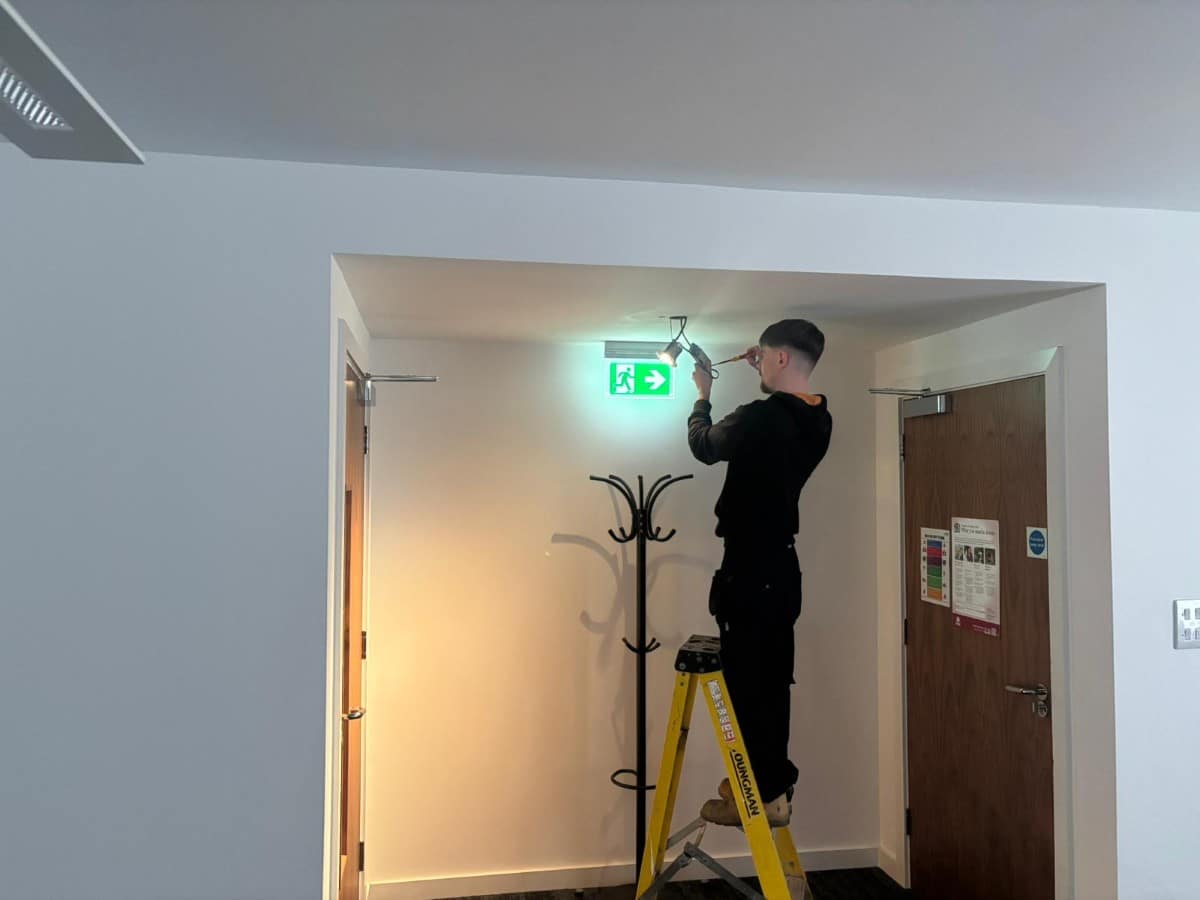 A person stands on a ladder fixing a light above an emergency exit sign in a hallway.