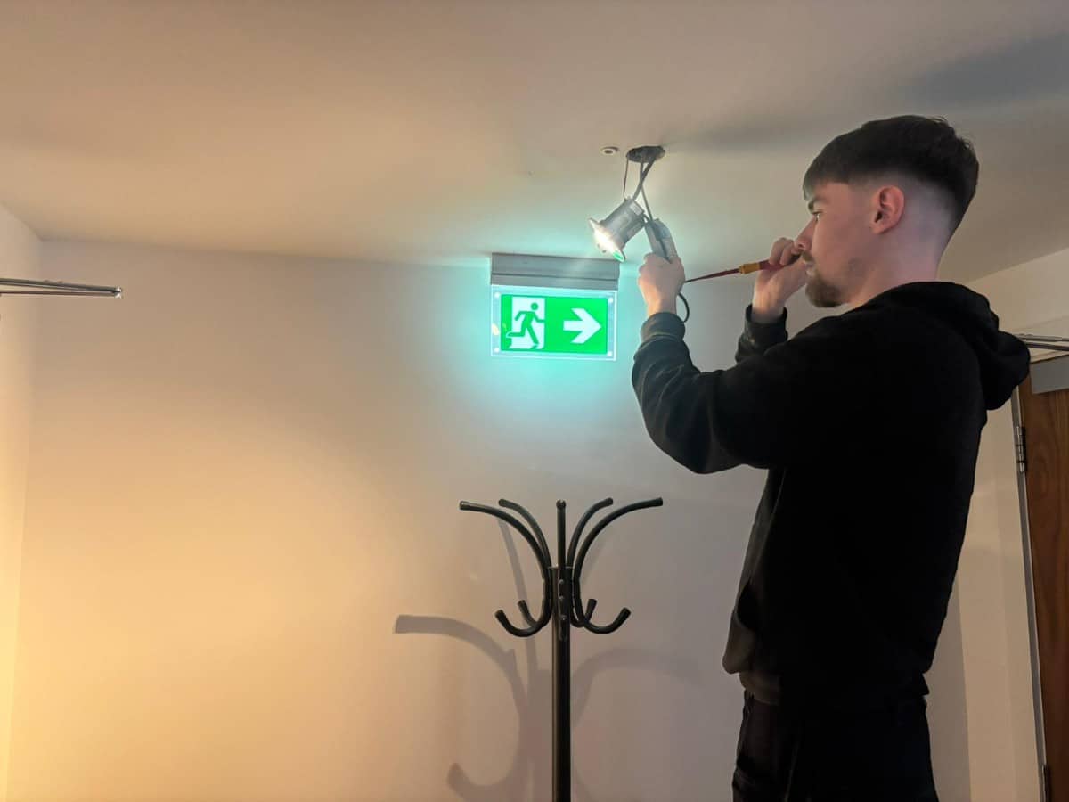 A man installs or checks an emergency light near an exit sign on the ceiling.