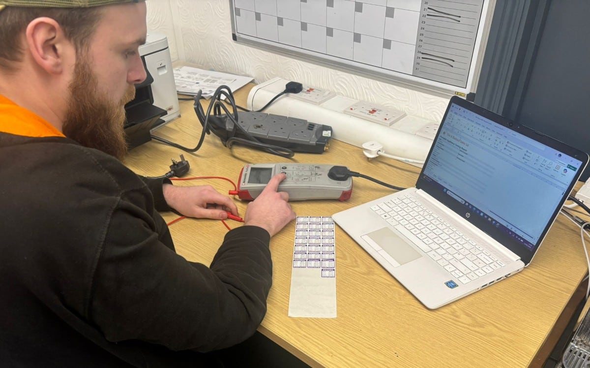 PAT Testing Done Right Man testing electrical equipment at a desk with a PAT tester, laptop, and power strip.