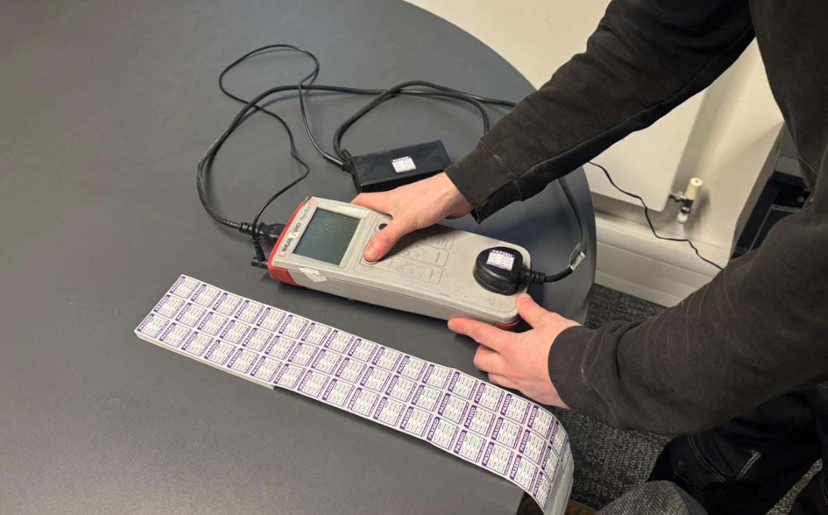 PAT Testing A person using a PAT testing device on a table, with a strip of test labels nearby.