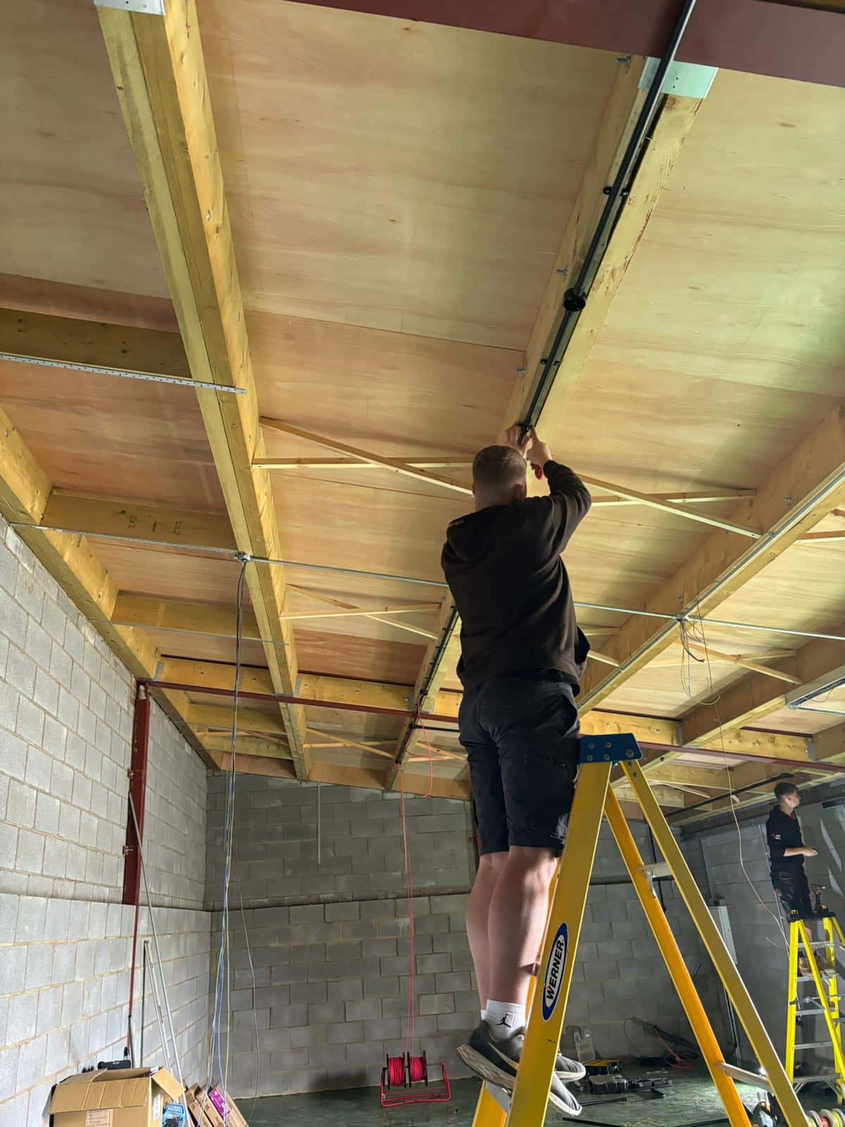 A person stands on a ladder fixing wiring to a ceiling in an unfinished room.