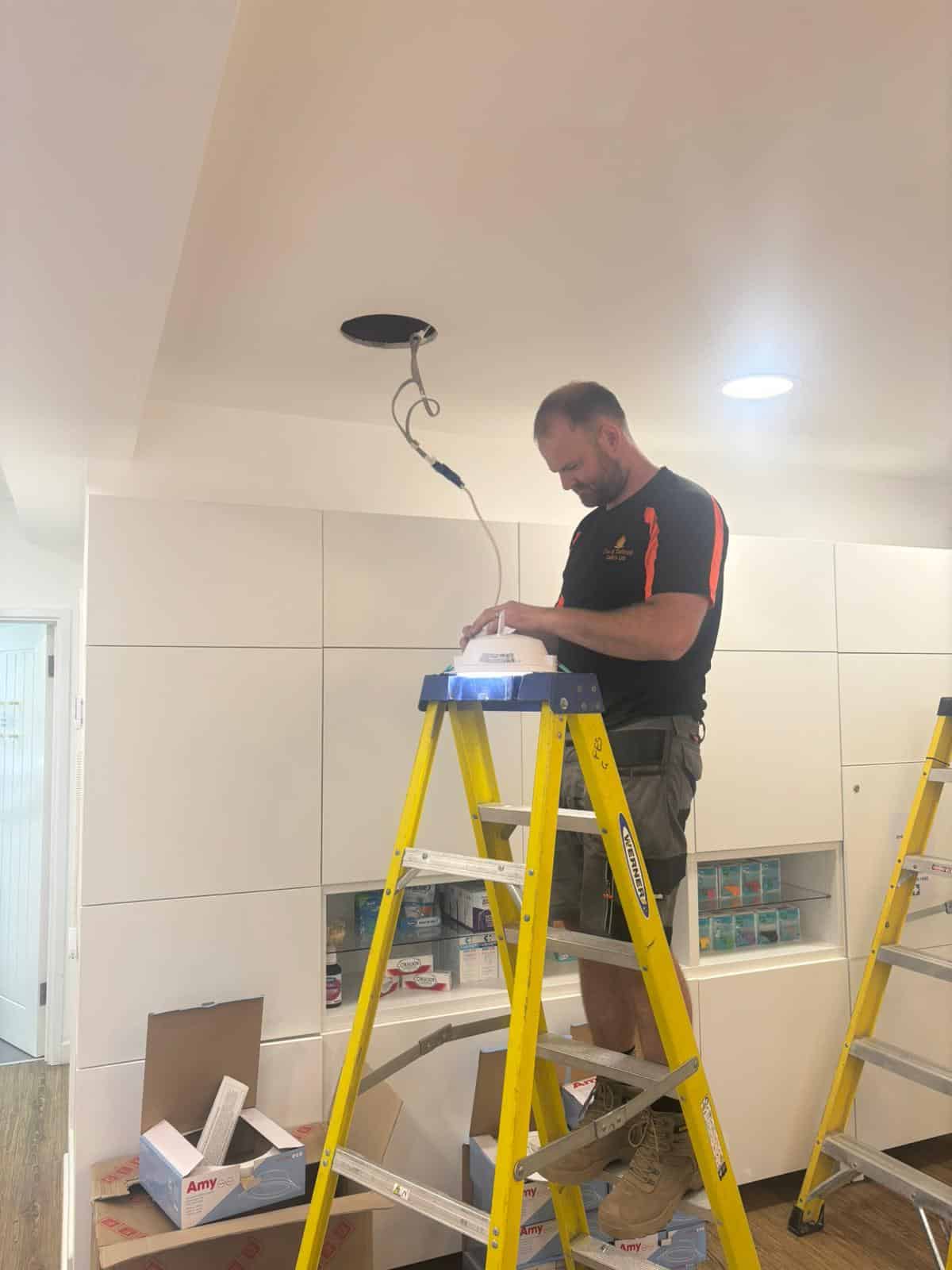 Man standing on a yellow ladder installing a light fixture in a ceiling.