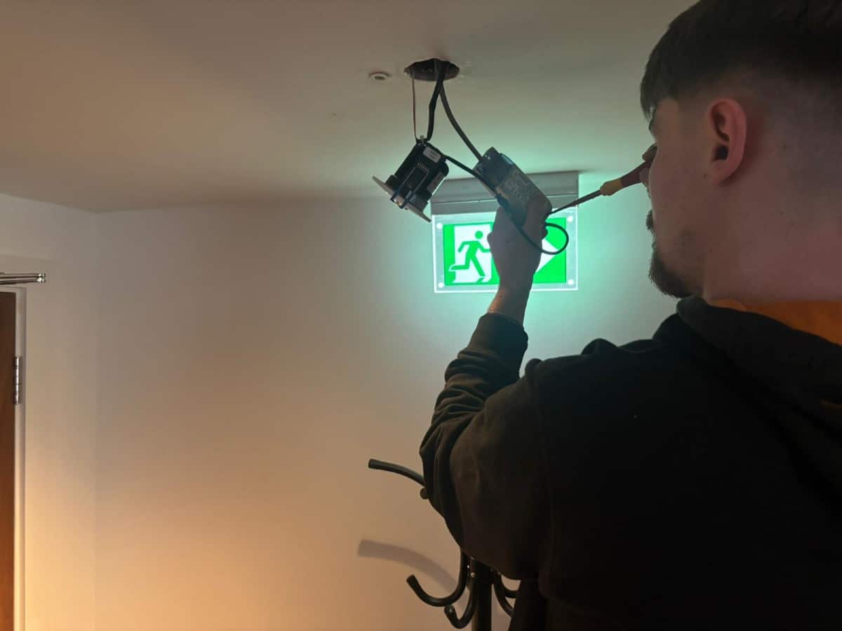 A person installs or repairs emergency lighting near an illuminated exit sign on a ceiling.